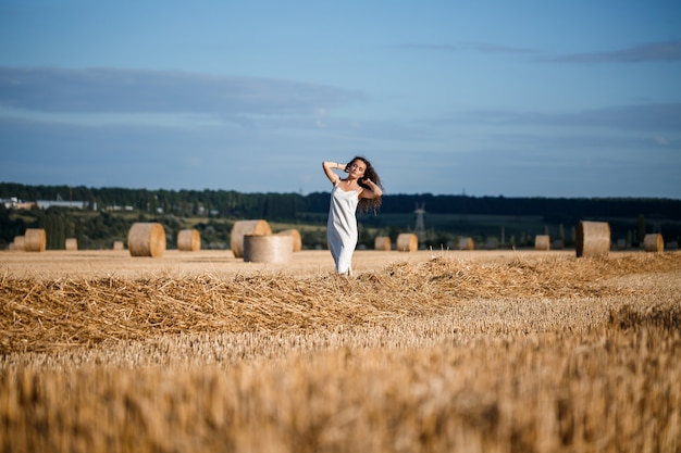 Young curly-haired woman in a wheat field, where there is a huge sheaf of hay, enjoying nature. People and travel. Nature. sun rays Agriculture