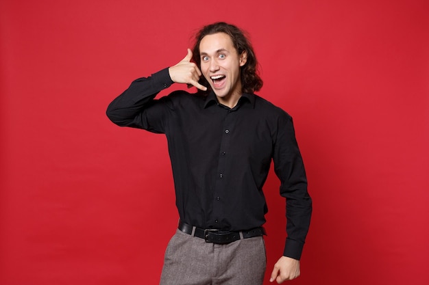 Young curly long haired man in black shirt posing isolated on red wall background studio portrait. People emotions lifestyle concept. Mock up copy space. doing phone gesture like says call me back.