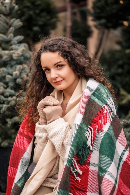 A young curly woman wrapped in a warm plaid in a cell sits waiting for the new year The girl on the background of green Christmas trees on the street Holiday mood