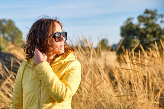 Young curlyhaired brownhaired girl in sunglasses and a yellow coat illuminated by the sunset sun