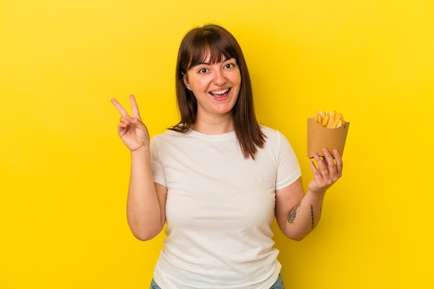 Young curvy caucasian woman holding fries isolated on yellow background joyful and carefree showing a peace symbol with fingers.