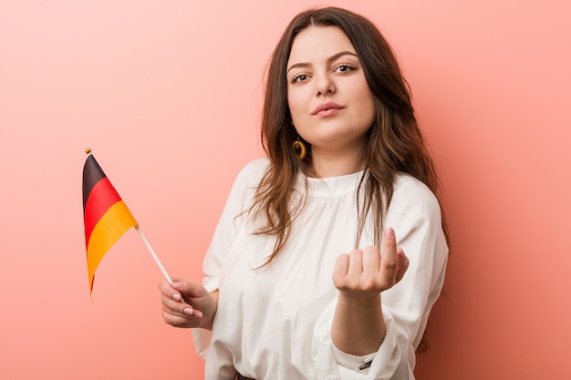 Photo young curvy plus size woman holding a germany flag pointing with finger at you as if inviting come closer.