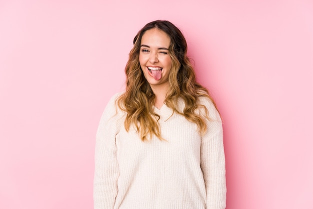 Young curvy woman posing in a pink wall isolated funny and friendly sticking out tongue.