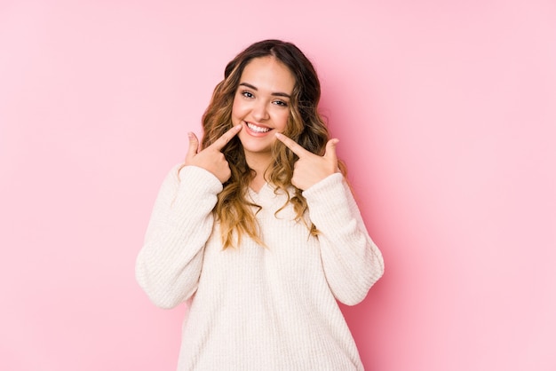 Young curvy woman posing in a pink wall isolated smiles, pointing fingers at mouth.