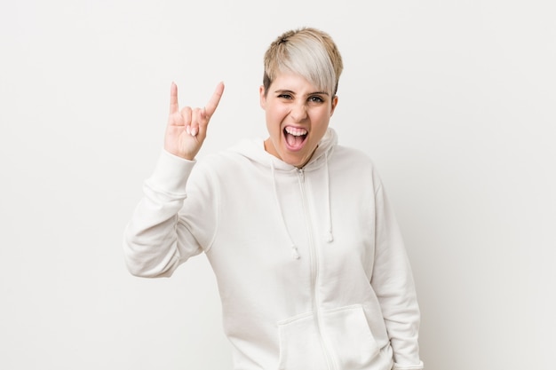 Young curvy woman wearing a white hoodie showing a horns gesture as a revolution concept.