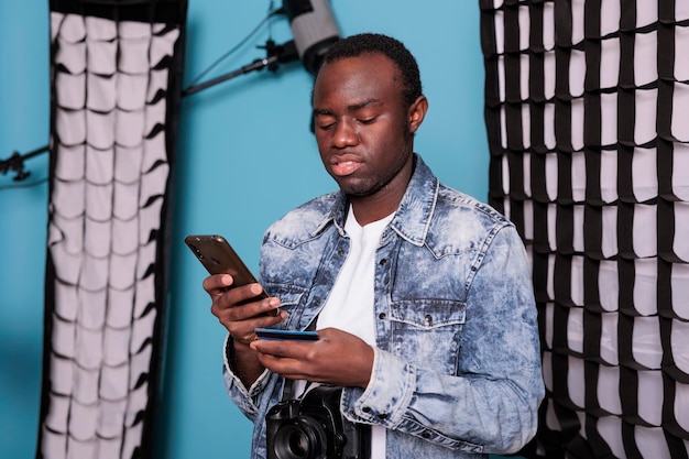 Young customer using debit card for online payment while standing in production studio with professional photo equipment. Man with DSLR camera paying cashless on digital retail store.