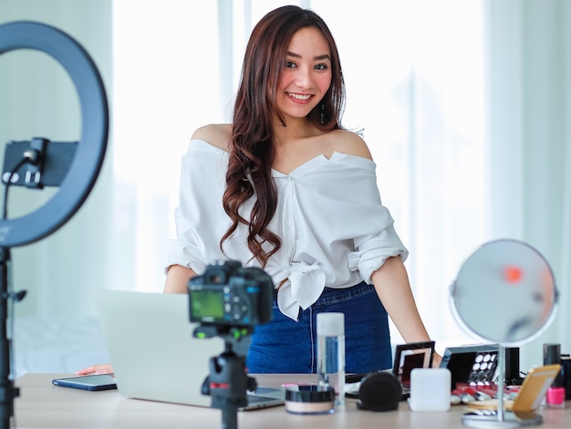 Young and cute Asian female vlogger, influencer, or online seller standing with cosmetics products and DSLR camera and notebook computer ready to broadcast online live stream or recording video.