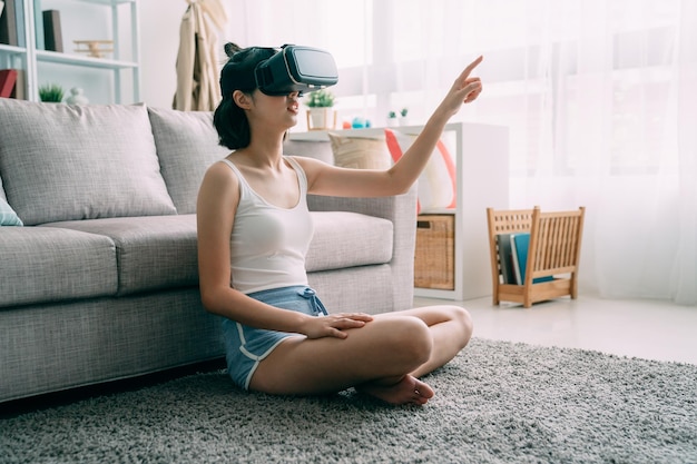 young cute asian japanese woman sit on carpet at home wearing virtual reality headset in home living room. charming girl in shorts in vr goggles point finger in house. lady on floor enjoy 3D glasses