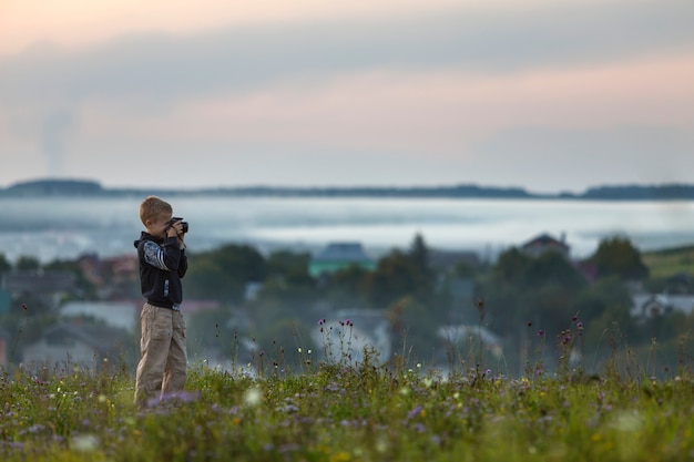 Photo young cute blond child boy with photo camera standing on grassy hill top on spring or summer day.