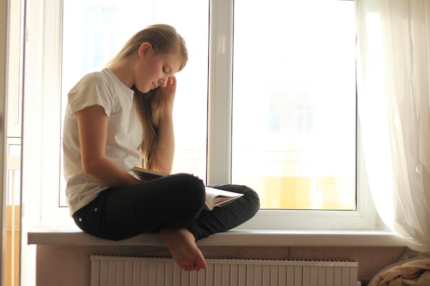 Young cute blonde teen girl sitting on windowsill and reading a book, youth hobby and leisure concept