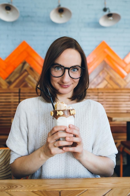 Young cute brunette woman drinks milkshake beautifully decorated with cream and cookies in the cafe. Toning.