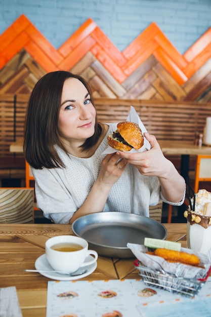 Young cute brunette woman eating a freshly prepared delicious Burger in the cafe. Toning.