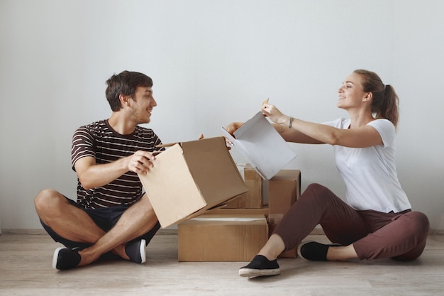 Young cute couple just married sitting on floor in a new apartment after repair among the cardboard boxes are happy and smiling.