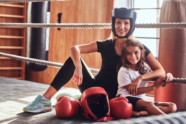 Young cute girl in helmet and her beautiful female boxing trainer are posing for photographer on the ring.