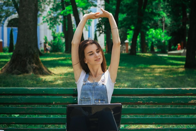 Young cute girl sitting with a laptop on the bench and raised her hands up