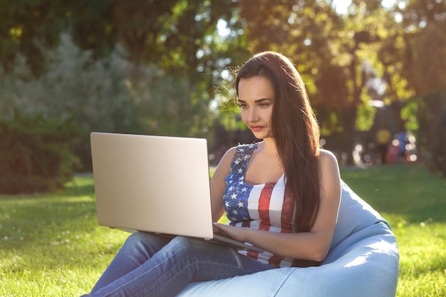 Young cute girl with laptop, sit on bean bag in garden or park, on green grass. Online shopping concept