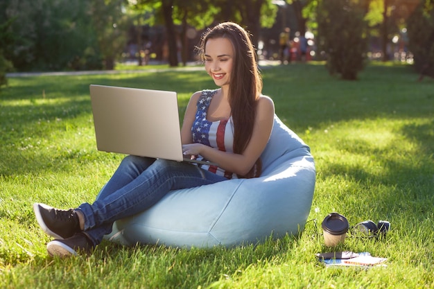 Young cute girl with laptop, sit on bean bag in garden or park, on green grass. Online shopping concept