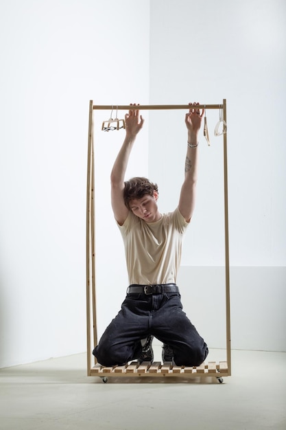 Young and cute guy in casual outfit posing on his knees with a wooden rail for hanging clothes