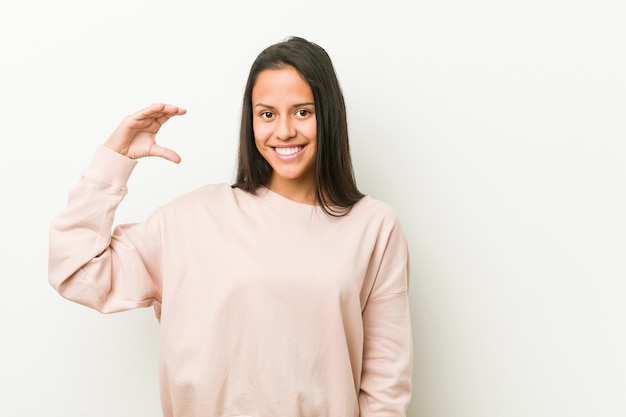 Young cute hispanic teenager woman holding something little with forefingers, smiling and confident.