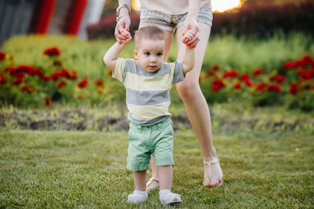 Photo a young cute mother helps and teaches her little son to take his first steps during sunset in the park on the grass.