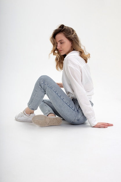 Young cute pensive caucasian girl posing in white shirt, blue jeans at studio