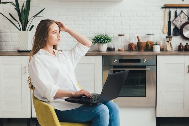 Young cute pregnant girl a freelancer working at home in the kitchen in the mode of self-isolation in quarantine