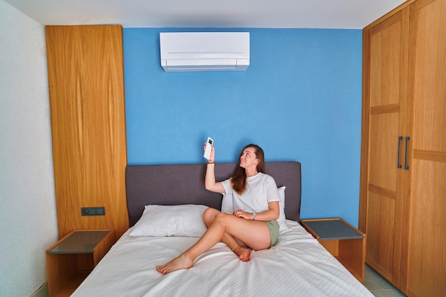 Young cute smiling girl lying on bed and adjusting temperature of air conditioner using remote control in bedroom