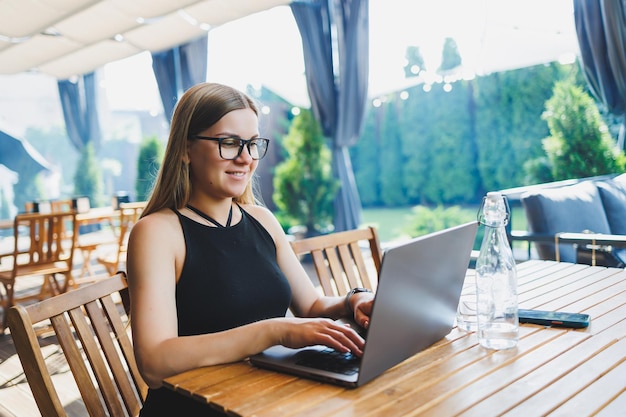 Young cute woman freelancer developer enjoying coffee break on summer terrace of cozy cafe working remotely on laptop smiling looking at laptop