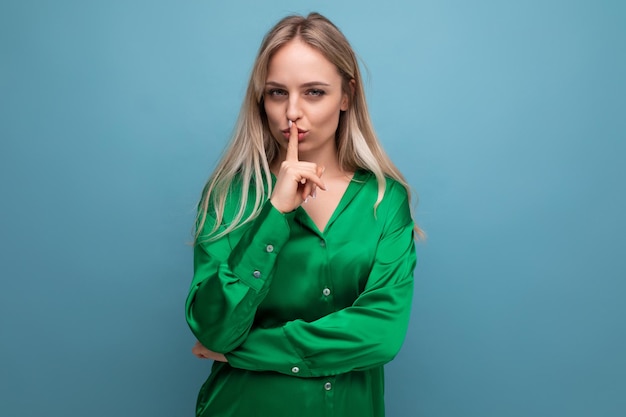 Young cute woman in a green shirt shows a secret with her finger on a blue studio background