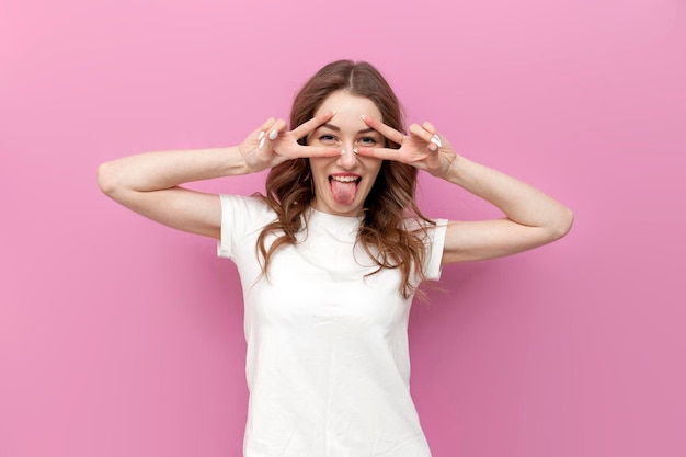 young cute woman shows tongue and makes peace gesture with hands on pink isolated background