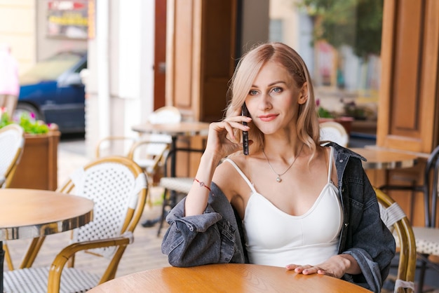 Young cute woman using phone sits in cafe at table with smartphone answering