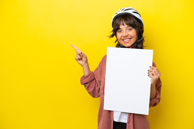 Young cyclist latin woman isolated on yellow background holding an empty placard and pointing side