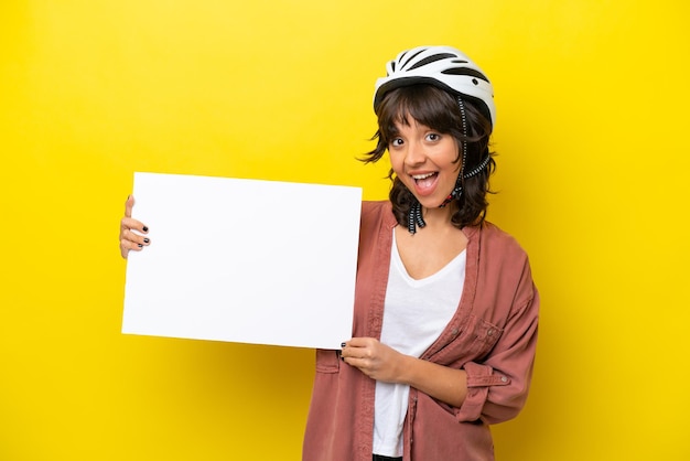 Young cyclist latin woman isolated on yellow background holding an empty placard with happy expression