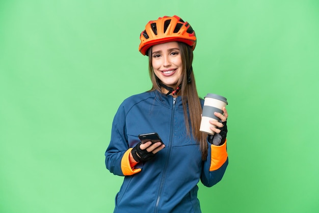 Young cyclist woman over isolated chroma key background holding coffee to take away and a mobile