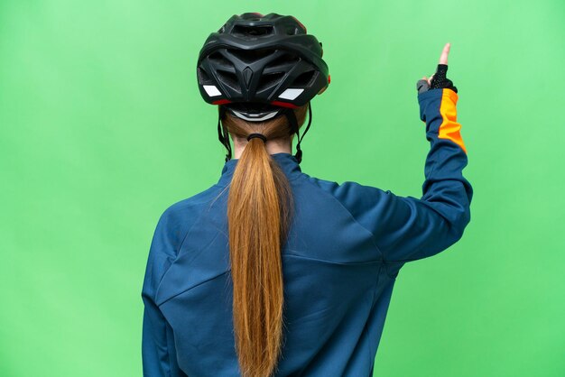 Young cyclist woman over isolated chroma key background pointing back with the index finger