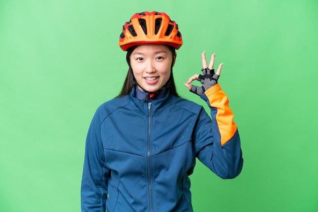 Young cyclist woman over isolated chroma key background showing ok sign with fingers