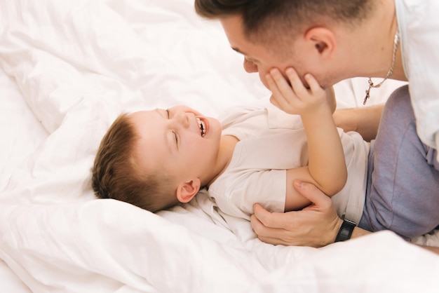 Young dad playing and laughing with his baby smiling son at home in bed in the bedroom happy family