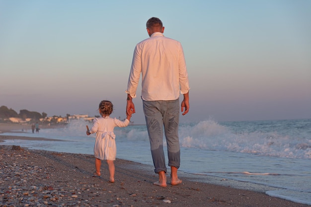 A young dad with a little daughter walks along the sea beach in the sunset