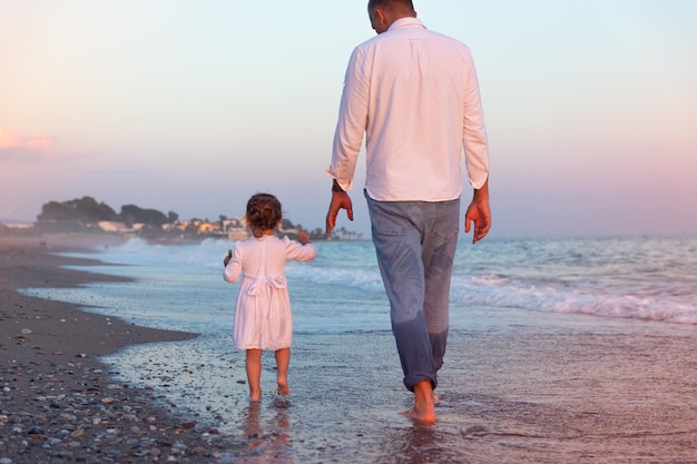 A young dad with a little daughter walks along the sea beach in the sunset