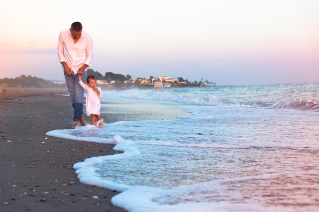 A young dad with a little daughter walks along the sea beach in the sunset