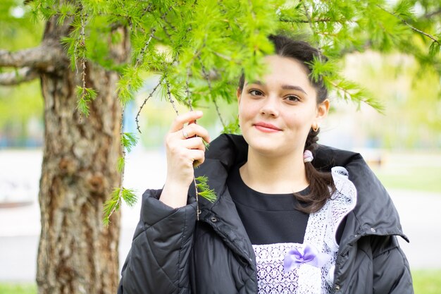 A young dark-haired girl on a background of green coniferous foliage. A smile on a pretty face.