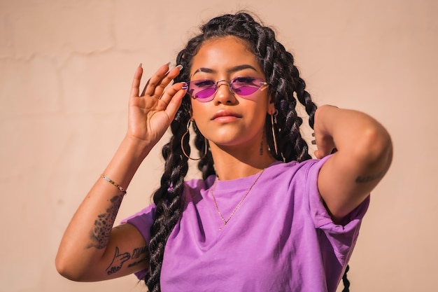  Young dark-skinned woman with long braids wearing purple glasses on a plain background, with serious look