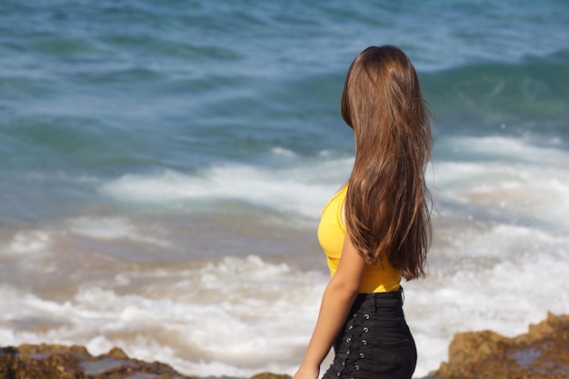 Young darkhaired beautiful European girl in yellow swimsuit smiling and standing on the background of the sea and splashes on a Sunny day