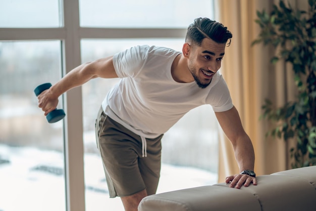 Young darkhaired man exercising with dumbbells and looking contented