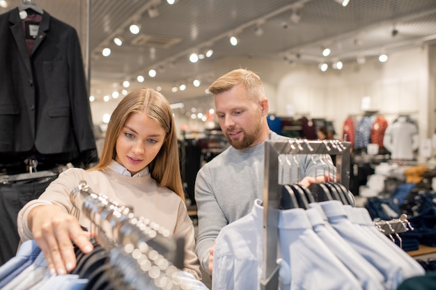 Young dates looking through collection of shirts hanging on racks while visiting large trade center at leisure