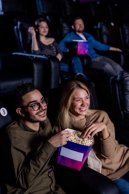 Young dates with box of popcorn sitting in dark cinema and looking at large screen while watching movie at leisure