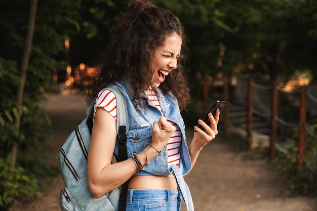 young delighted woman wearing backpack, clenching fist and screaming with smartphone in hand while walking through green park