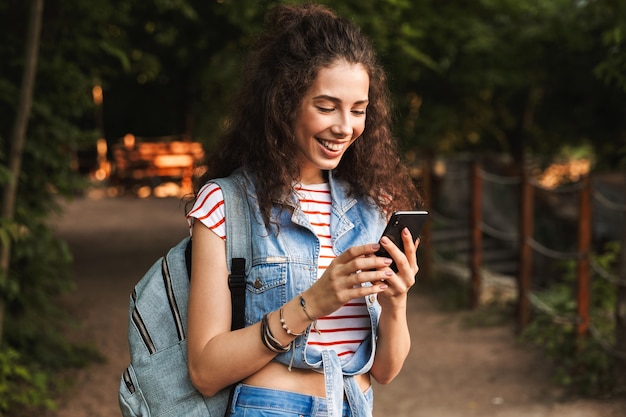 young delighted woman with backpack, smiling and looking at smartphone in hand while standing on path in green park