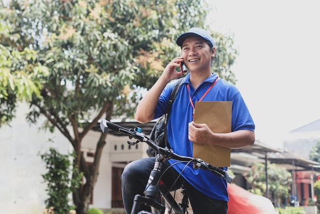 Young delivery man holding brown envelope having a call on bicycle