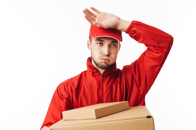 Young delivery man in red cap and jacket holding boxes in hands tiredly 